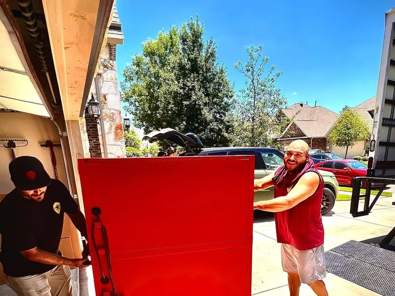 Two movers rolling a heavy red cabinet out of a garage on a dolly